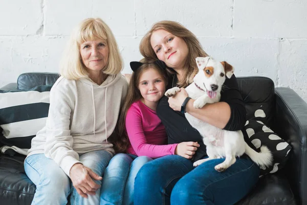 Happy granddaughter and grandmother and daughter sitting on sofa with jack russell terrier dog. Grandma hugs grandchild at home. Relationship, family and three generation concept. — Stock Photo, Image