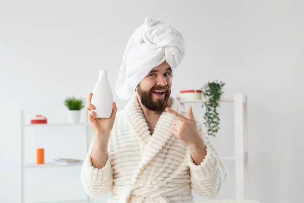 Beauty portrait of handsome beautiful guy in towel and bathrobe holding tube of body cream lotion in hand. Spa, body and skin care for man concept