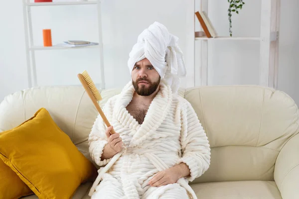 Retrato de cara alegre usar toalha de turbante segurando em mãos escova de massagem enquanto se senta no sofá. Cuidados com a pele masculina e conceito spa. — Fotografia de Stock