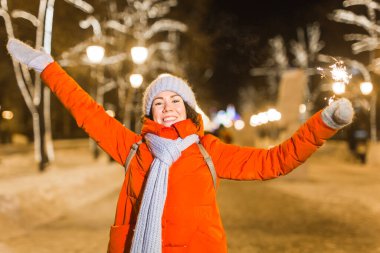 Kış örgü giysileri giymiş, dışarıda kar altında maytap tutan gülümseyen genç bir kadın. Noel tatili.