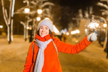 Kış örgü giysileri giymiş, dışarıda kar altında maytap tutan gülümseyen genç bir kadın. Noel tatili.