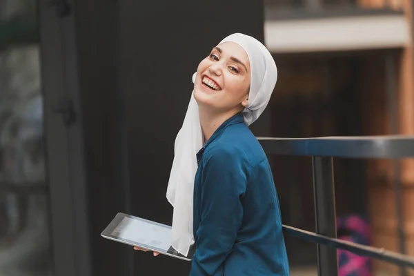 Улыбающаяся мусульманка с помощью цифрового планшета, просмотра Интернета или социальных сетей на открытом воздухе — стоковое фото