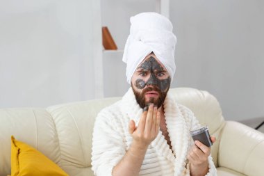 Sakallı adamın cildi temiz, yüzüne kil maske takıyor ve güzellik tedavisinden hoşlanıyor. Evde spa, vücut ve cilt bakımı