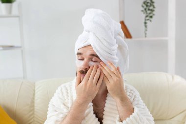 Evde gözlerinin altında yamalar olan genç bir adam. Erkekler için kozmetoloji kavramı. Evde spa, vücut ve cilt bakımı