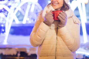Noel tatili. Kışın sıcak bir fincan kahveyle sıcak kıyafetler içinde gülümseyen güzel bir kadın. Şehir gecesi.