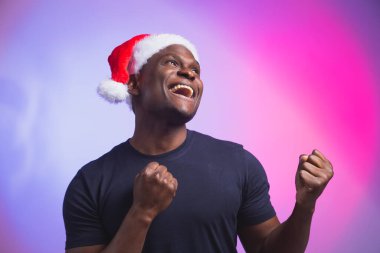 Noel Baba şapkalı ve renkli arka planda sıradan tişörtlü Afro-Amerikan gülümseyen adam portresi. Noel zamanı. Kış tatilleri ve duygusal insanlar konsepti.