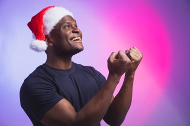 Noel Baba şapkalı ve renkli arka planda sıradan tişörtlü Afro-Amerikan gülümseyen adam portresi. Noel zamanı. Kış tatilleri ve duygusal insanlar konsepti.