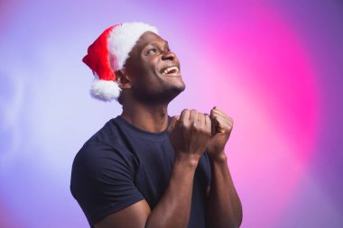 Noel Baba şapkalı ve renkli arka planda sıradan tişörtlü Afro-Amerikan gülümseyen adam portresi. Noel zamanı. Kış tatilleri ve duygusal insanlar konsepti.