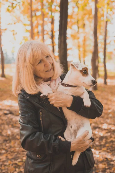 Mujer sonriente mayor abrazando a su perro en el parque de otoño. Envejecimiento activo y concepto de mascota. — Foto de Stock