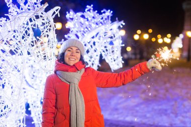 Kış örgü giysileri giymiş, dışarıda kar altında maytap tutan gülümseyen genç bir kadın. Noel tatili.
