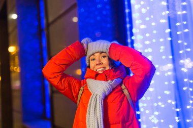 Noel dekorasyon ışıkları sokağında eğlenen bir kız. Açık havada şık örgü örüp ceket giyen mutlu, genç bir kadın. Model gülüşü. Kış Harikalar Diyarı Sahnesi, Yeni Yıl Partisi.