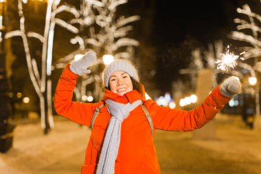 Kış örgü giysileri giymiş, dışarıda kar altında maytap tutan gülümseyen genç bir kadın. Noel tatili.