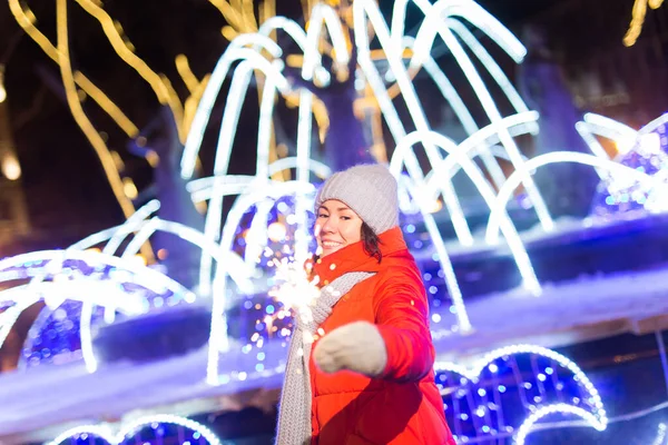 Giovane donna sorridente che indossa vestiti a maglia invernali che tengono scintillante all'aperto su sfondo neve. Vacanze di Natale. — Foto Stock