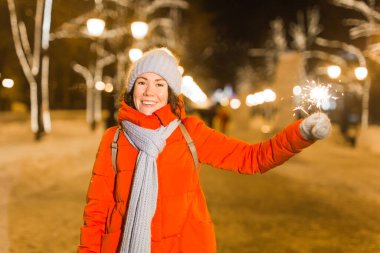 Kış örgü giysileri giymiş, dışarıda kar altında maytap tutan gülümseyen genç bir kadın. Noel tatili.