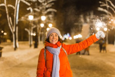 Kış örgü giysileri giymiş, dışarıda kar altında maytap tutan gülümseyen genç bir kadın. Noel tatili.
