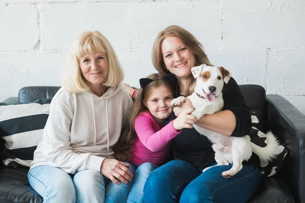 Glad barnbarn och mormor och dotter sitter på soffan med jack russell terrier hund. Mormor kramar barnbarn hemma. Förhållande, familj och tre generationer. — Stockfoto