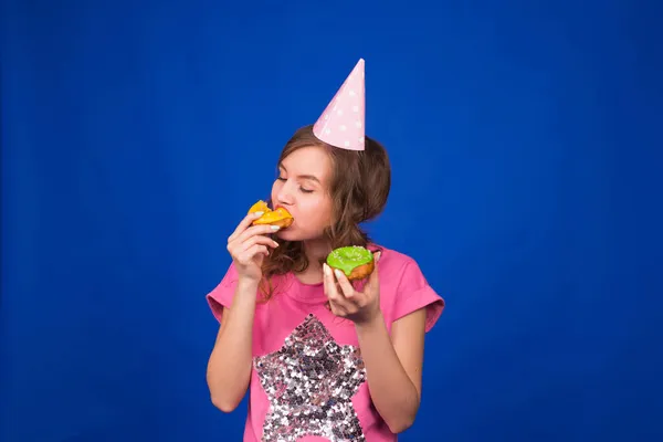 Μια νεαρή όμορφη γυναίκα τρώει ντόνατς σε μπλε φόντο. Ανθυγιεινή διατροφή, πρόχειρο φαγητό και εθισμός στα τρόφιμα — Φωτογραφία Αρχείου