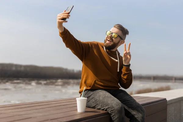 Heyecanlı genç adam akıllı telefondan selfie çekiyor. Z jenerasyonunda ya da milenyumda ve bahar kasabasında yürüyüş yaparken günlük moda kıyafetleri giyiyor. Yaşam tarzı kavramı — Stok fotoğraf