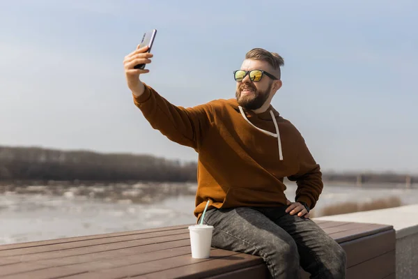 Heyecanlı genç adam akıllı telefondan selfie çekiyor. Z jenerasyonunda ya da milenyumda ve bahar kasabasında yürüyüş yaparken günlük moda kıyafetleri giyiyor. Yaşam tarzı kavramı — Stok fotoğraf