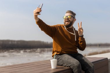 Sakallı adam baharın tadını çıkarıyor ve akıllı telefon kullanarak gezinti alanında selfie çekiyor. Teknoloji boş zaman ve gen z konsepti