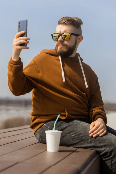 Νεαρός hipster άνθρωπος χρησιμοποιώντας έξυπνο τηλέφωνο κάθεται σε ξύλινες σανίδες. Επικοινωνία στα μέσα κοινωνικής δικτύωσης — Φωτογραφία Αρχείου
