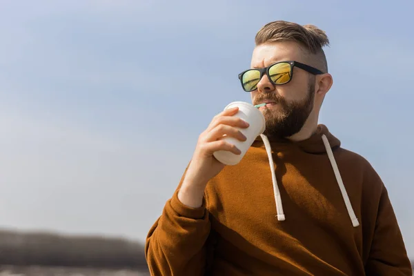 Een man met baard die een milkshake drinkt in de voorjaarspromenade. Drankjes weg te nemen en gen z of millennial jeugdconcept. Kopieerruimte voor reclame — Stockfoto