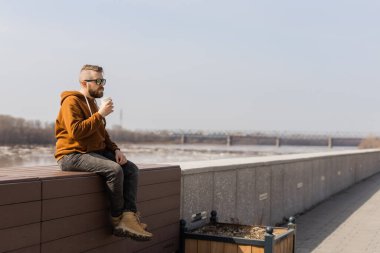 Sakallı düşünceli adam içki içerek uzaklaşıyor. Nehir kıyısına ve gökyüzüne bakıyor. Arka planda fotokopi alanı var.