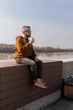 Sakallı, düşünceli adam içki içerken nehir kıyısına ve gökyüzüne bakıyor. Çekici, yalnız erkek, şehrin arka planında oturuyor.