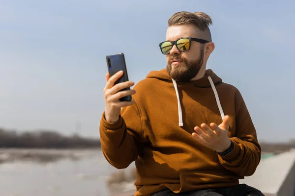 Тысячелетний бородатый мужчина делает видеозвонок по мобильному телефону на открытом воздухе в городе. Концепция смартфонов и технологий — стоковое фото
