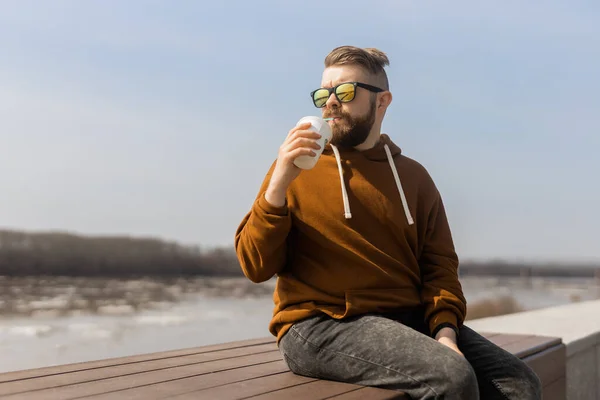 Hombre barbudo bebiendo un batido en el paseo marítimo de primavera. Bebidas para llevar y gen z o concepto de juventud milenaria. Espacio de copia para publicidad — Foto de Stock