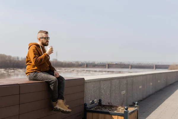 Bearded thoughtful man holds drink looking away while standing against river shore and sky background with copy space area for your text , attractive lonely male sits on city promenade background — Stock Photo, Image