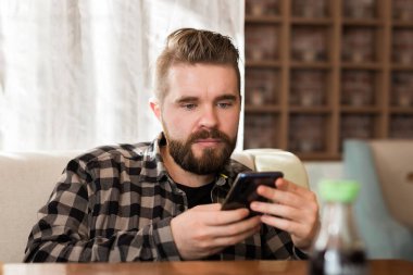 Modern kahve dükkanında akıllı telefon kullanan gülümseyen bir erkek, cep telefonunda mesajlaşarak sohbet ediyor. Sosyal ağlar ve milenyum konsepti
