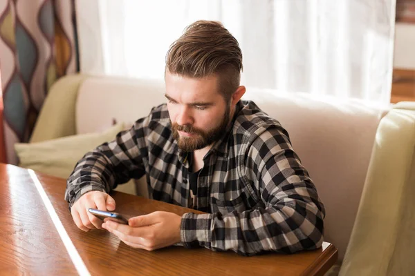 Joven chico hipster buscando noticias de las redes sociales leer a través de teléfono inteligente en tiempo libre en la cafetería — Foto de Stock