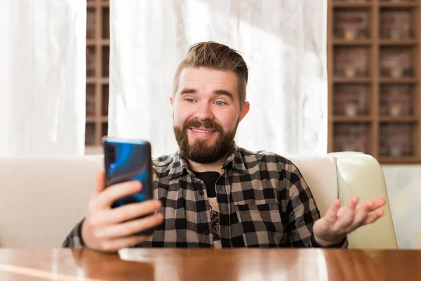 Feliz joven sentado en la toma de café con auriculares haciendo videollamada a un amigo. Caucásico macho tener vídeo chat en el teléfono inteligente en la cafetería. — Foto de Stock