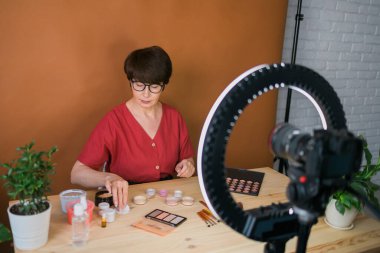 Middle-aged woman talking on cosmetics with makeup eye shadows and blush palette and brushes while recording her video. Mature female making video for her blog on cosmetics