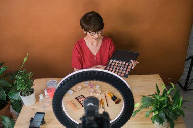Middle-aged woman talking on cosmetics with makeup eye shadows and blush palette and brushes while recording her video. Mature female making video for her blog on cosmetics