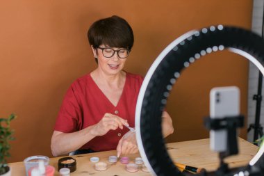 Middle-aged woman talking on cosmetics with makeup eye shadows and blush palette and brushes while recording her video. Mature female making video for her blog on cosmetics