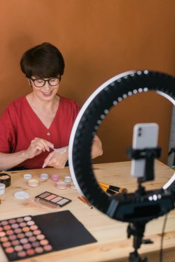 Beauty and cosmetics e-commerce and blogging concept - professional mature female make up artist testing new cosmetics recording video review on cosmetic products at home
