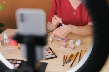 Close-up Middle-aged female beauty blogger or influencer with makeup product recording video in room at home. Using ring lamp and smartphone