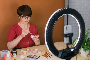 Middle-aged woman talking on cosmetics with makeup eye shadows and blush palette and brushes while recording her video. Mature female making video for her blog on cosmetics