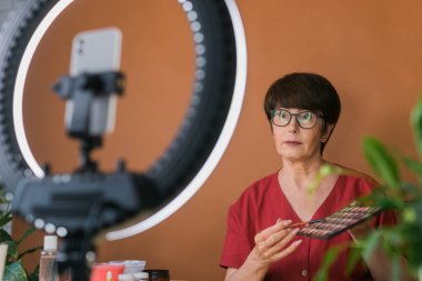 Middle-aged woman talking on cosmetics with makeup eye shadows and blush palette and brushes while recording her video. Mature female making video for her blog on cosmetics