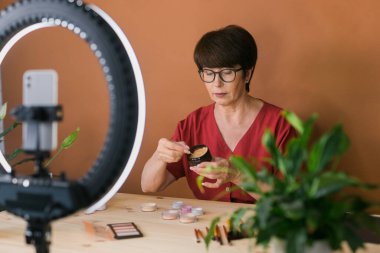 Beauty and cosmetics e-commerce and blogging concept - professional mature female make up artist testing new cosmetics recording video review on cosmetic products at home