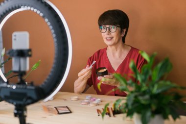 Middle-aged female beauty blogger or influencer with makeup product recording video in room at home. Using ring lamp and smartphone