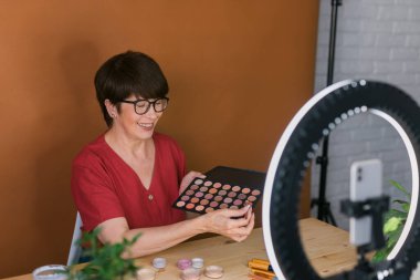 Beauty and cosmetics e-commerce and blogging concept - professional mature female make up artist testing new cosmetics recording video review on cosmetic products at home