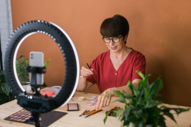 Middle-aged woman talking on cosmetics with makeup eye shadows and blush palette and brushes while recording her video. Mature female making video for her blog on cosmetics