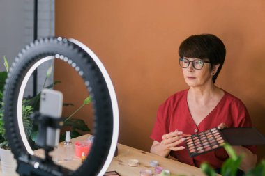 Middle-aged female beauty blogger or influencer with makeup product recording video in room at home. Using ring lamp and smartphone