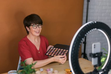 Beauty and cosmetics e-commerce and blogging concept - professional mature female make up artist testing new cosmetics recording video review on cosmetic products at home