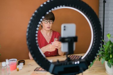 Middle-aged woman talking on cosmetics with makeup eye shadows and blush palette and brushes while recording her video. Mature female making video for her blog on cosmetics