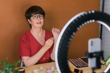 Beauty and cosmetics e-commerce and blogging concept - professional mature female make up artist testing new cosmetics recording video review on cosmetic products at home