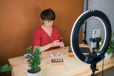 Middle-aged woman talking on cosmetics with makeup eye shadows and blush palette and brushes while recording her video. Mature female making video for her blog on cosmetics